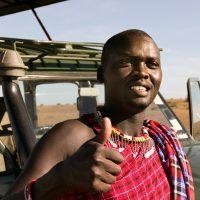 Maasai guide leaning against safari jeep and giving a thumbs up.