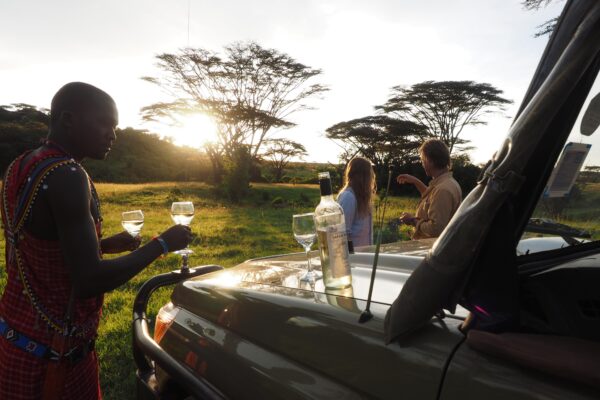 Couple on a Basecamp Explorer game drive, looking at sunset on the savanna in Masai Mara, Maasai guide holding wineglasses, bottle of white wine standing on the hood of the car.
